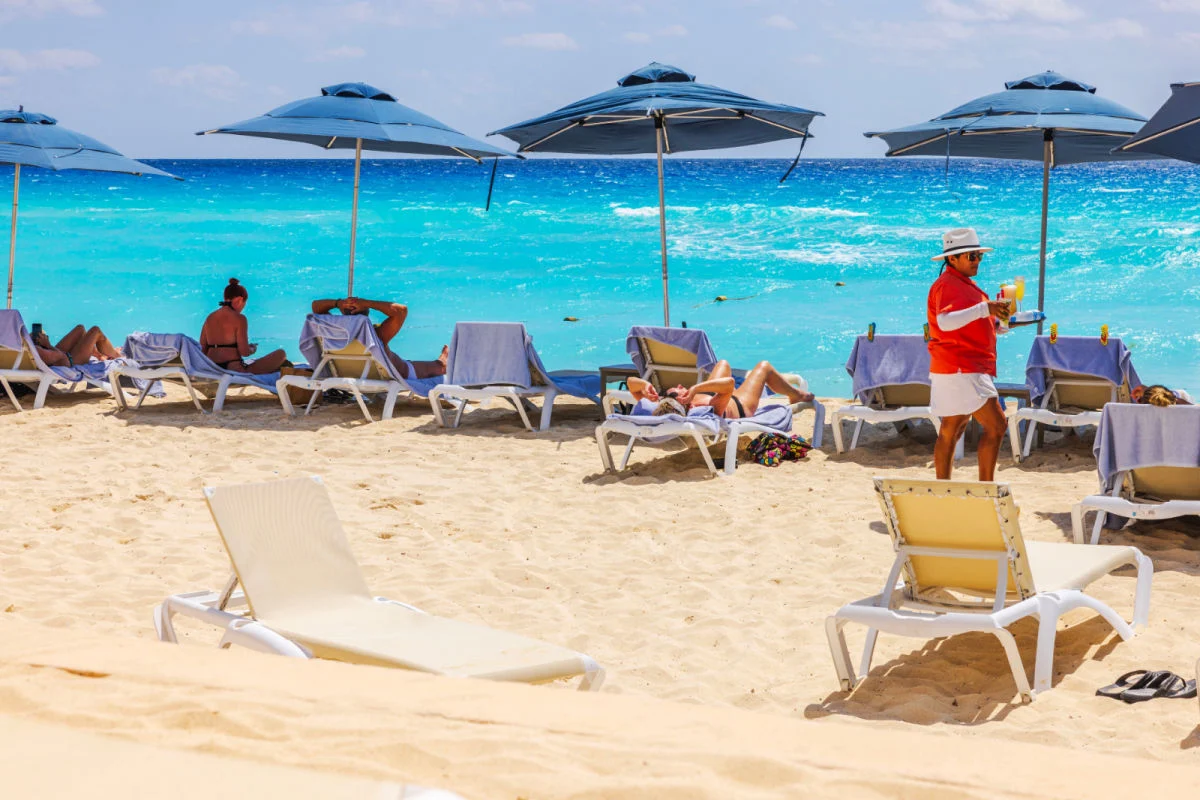 Plage-des-Caraïbes-avec-des-touristes-se-relaxant-sur-des-chaises-longues-sous-des-parapluies-bleus-pendant-que-le-serveur-sert-des-boissons-contre-les-vagues-turquoises.-Mexique.-Cancun