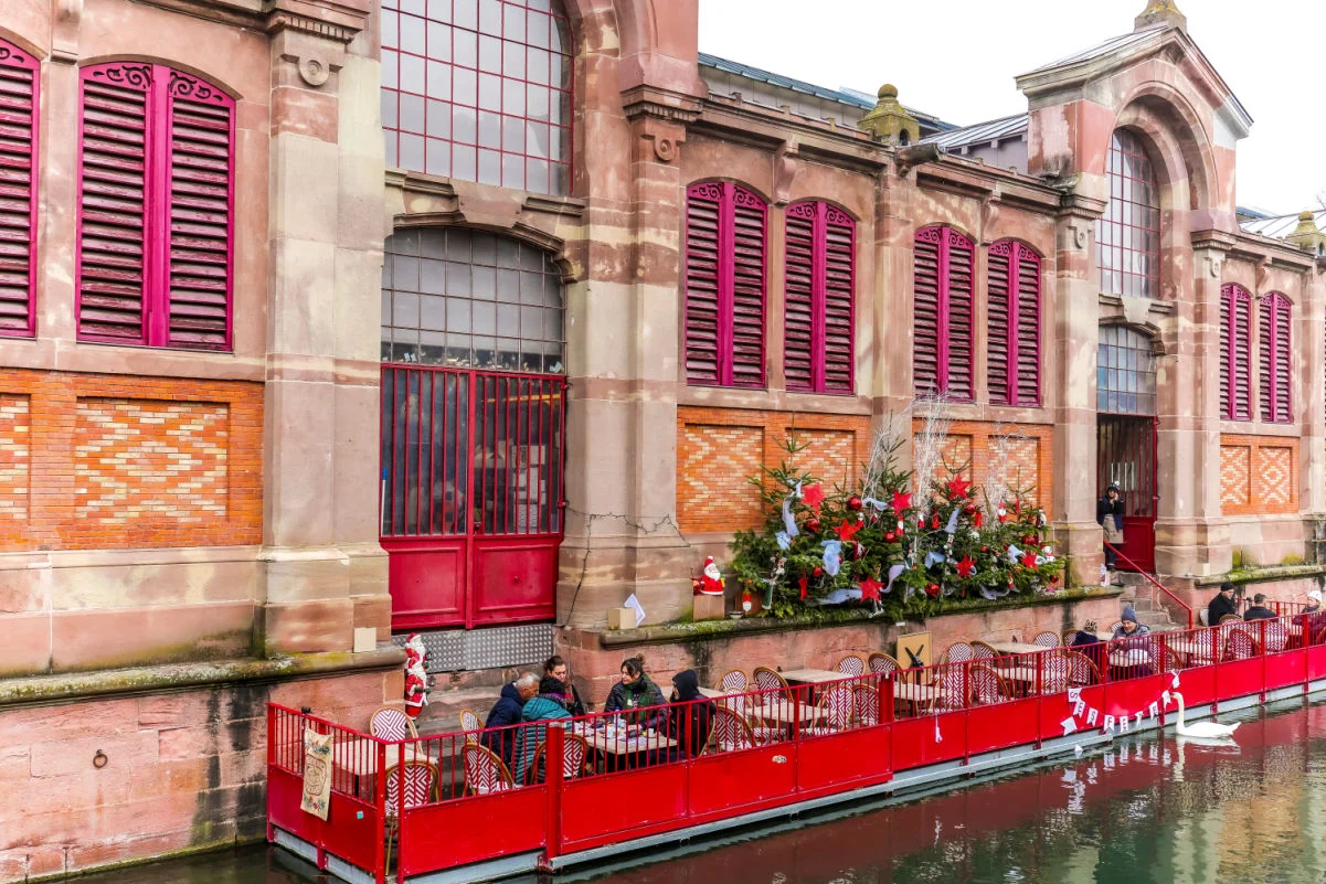 Les gens dînent dans un café avec terrasse au bord de la rivière, à l'extérieur du bâtiment historique Marche Couvert (marché couvert) dans le quartier de la Petite Venise de Colmar, en Alsace, pendant la saison de Noël.